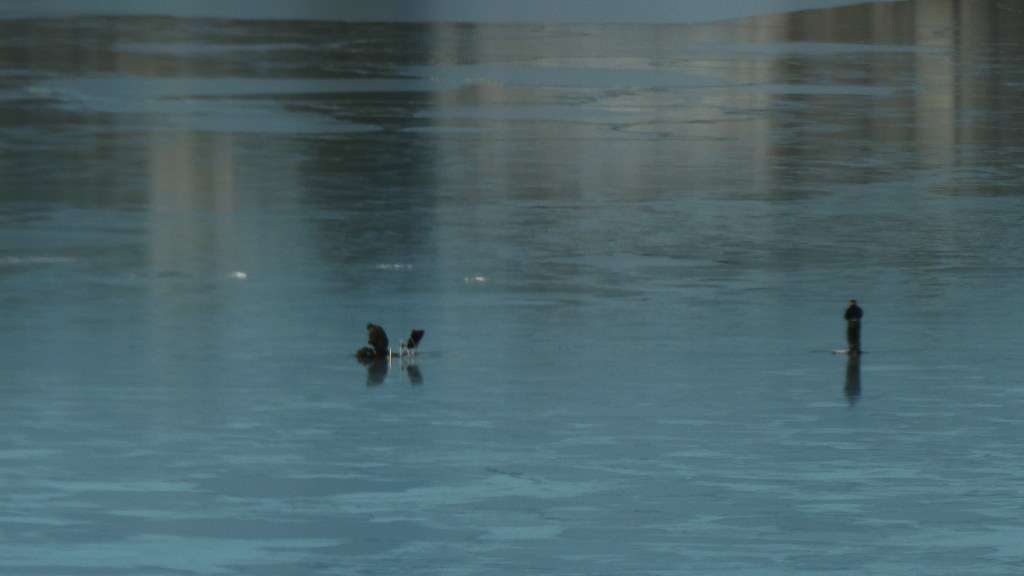 To isfiskere på isen, tatt med telelinse fra stuen min. Du ser den blå stålisen og én fisker til høyre i bildet som står oppreist ved en råk, og én i midten av bildet som sitter ved en sammenleggbar campingstol ved en annen råk.