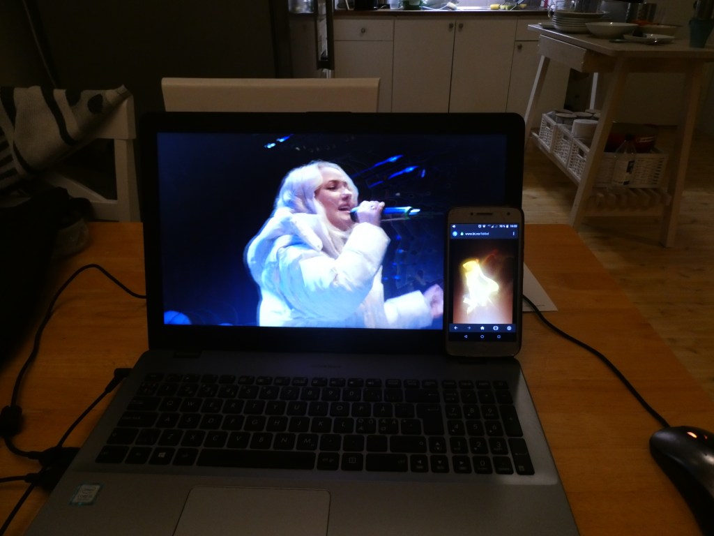 Laptopen min på kjøkkenbordet, livestream av lysfesten. Mobilen min står inntil PC-skjermen og viser fakkelflamme.